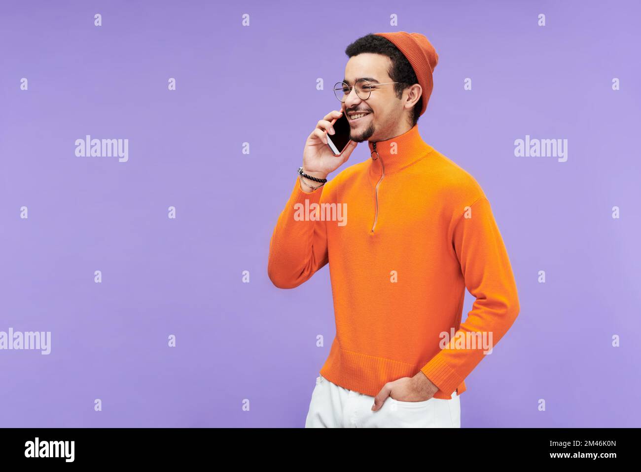 Hombre joven feliz en cuello alto, gorro y anteojos que se comunican en el teléfono inteligente mientras se coloca delante de la cámara Foto de stock
