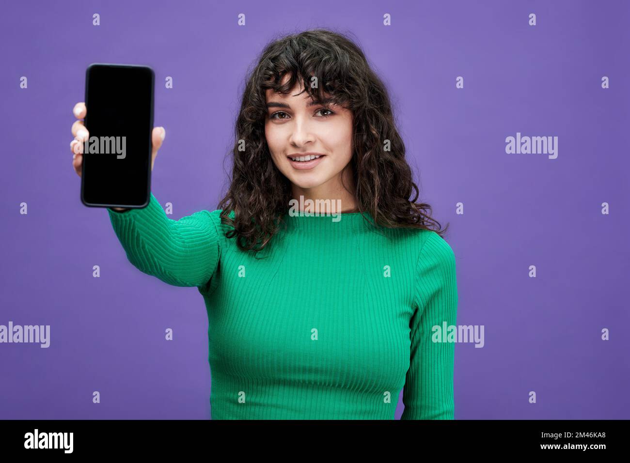 Chica morena en pullover verde con mangas largas sosteniendo smartphone mientras hace la presentación del anuncio de venta sobre fondo violeta Foto de stock
