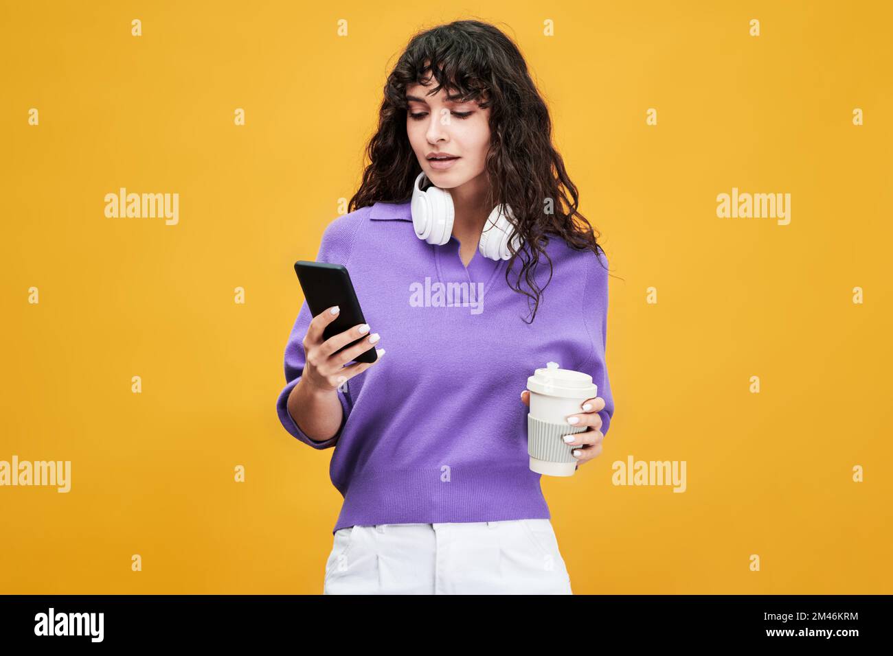 Estudiante femenino lindo en v-camisa de lavanda y mientras que pantalones usando el teléfono móvil y teniendo taza de té en el descanso contra el fondo amarillo Foto de stock