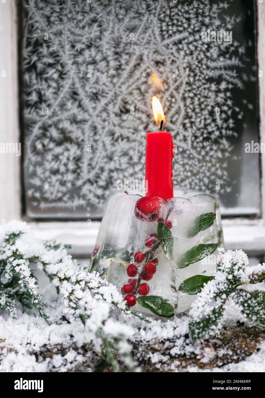Candelabro de Navidad hecho a mano en forma de pastel hecho de hielo, ramas de bayas de acebo rojo con vela ardiente frente a la ventana cubierta de escarcha. Foto de stock