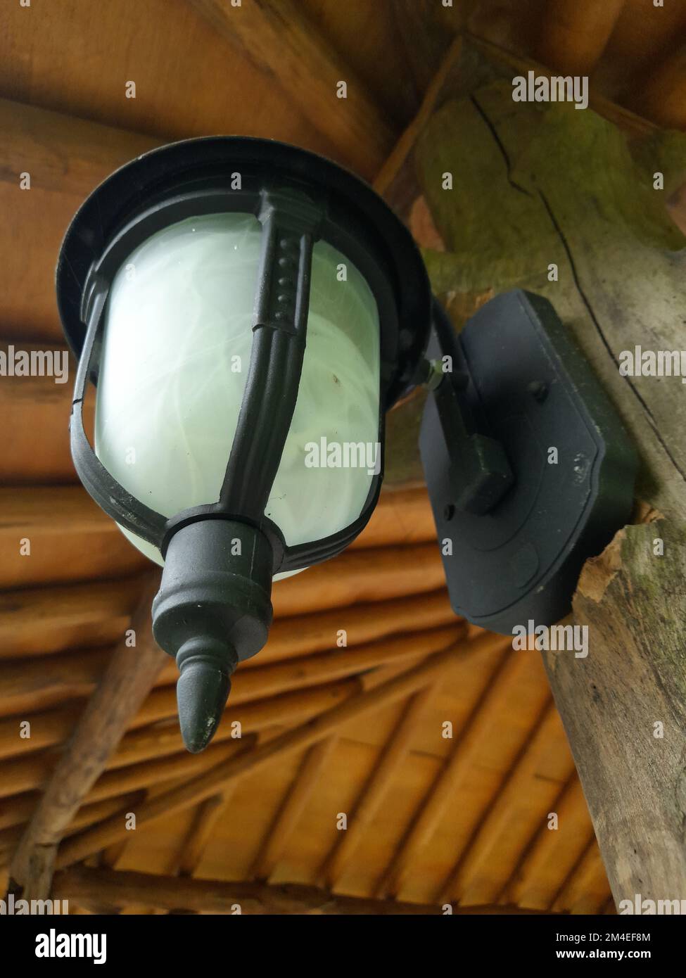 Lámpara de jardín antigua y minimalista con espejo blanco y tallo negro unido a un poste de madera contra un fondo de techo de madera Foto de stock