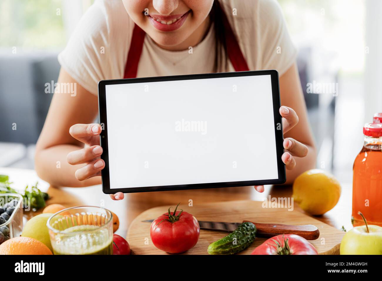 Manos de la mujer que sostienen la tableta, mostrando la maqueta blanca vacía de la pantalla en blanco en la cocina mientras que cocina el desayuno vegetal saludable. Compras de alimentos en línea. Entrega de anuncios de aplicaciones, libro de cocina, recetas de plan de dieta Foto de stock