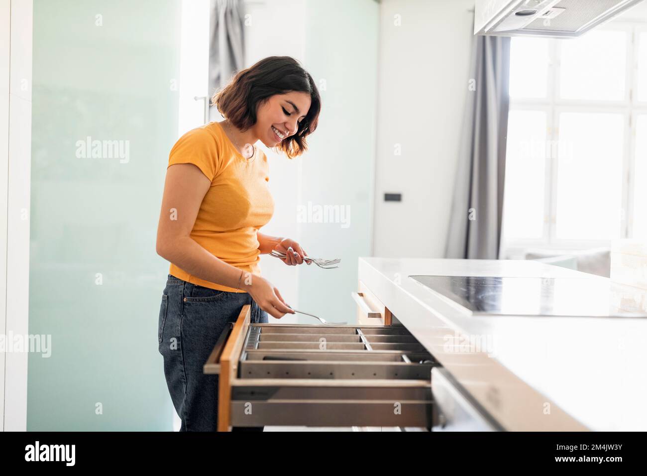 Mujer sonriente del Medio Oriente ordenando hasta los cubiertos en cajón de la cocina, vista lateral Foto de stock
