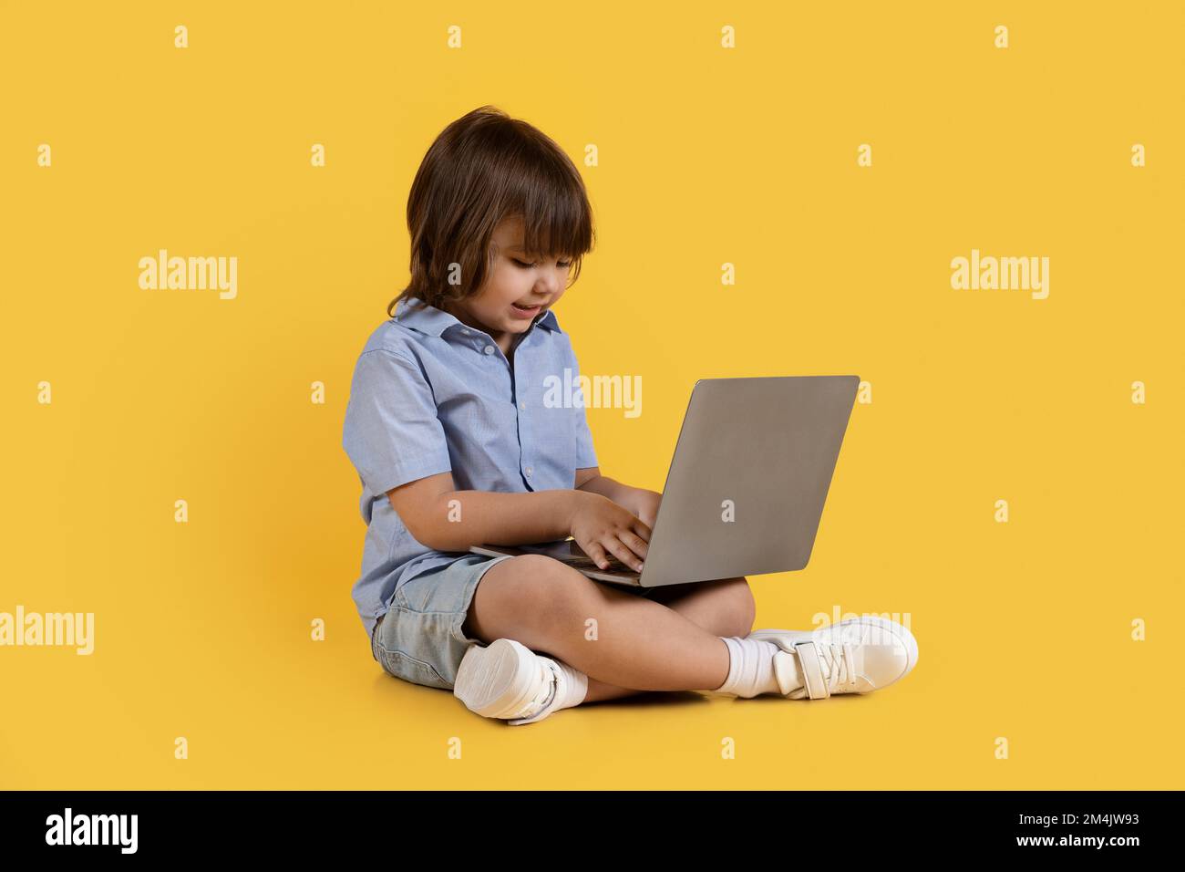 Adorable niño pequeño escribiendo en la computadora portátil, jugando a desarrollar el juego, sentado en el piso, fondo de estudio naranja Foto de stock