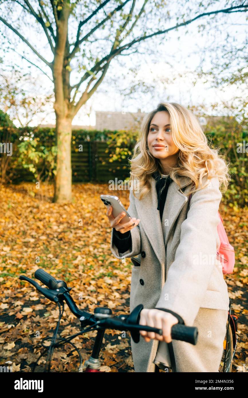 Mujer caucásica atractiva joven que usa el teléfono móvil, paseo en bicicleta en el parque en día cálido en el otoño. Foto de stock