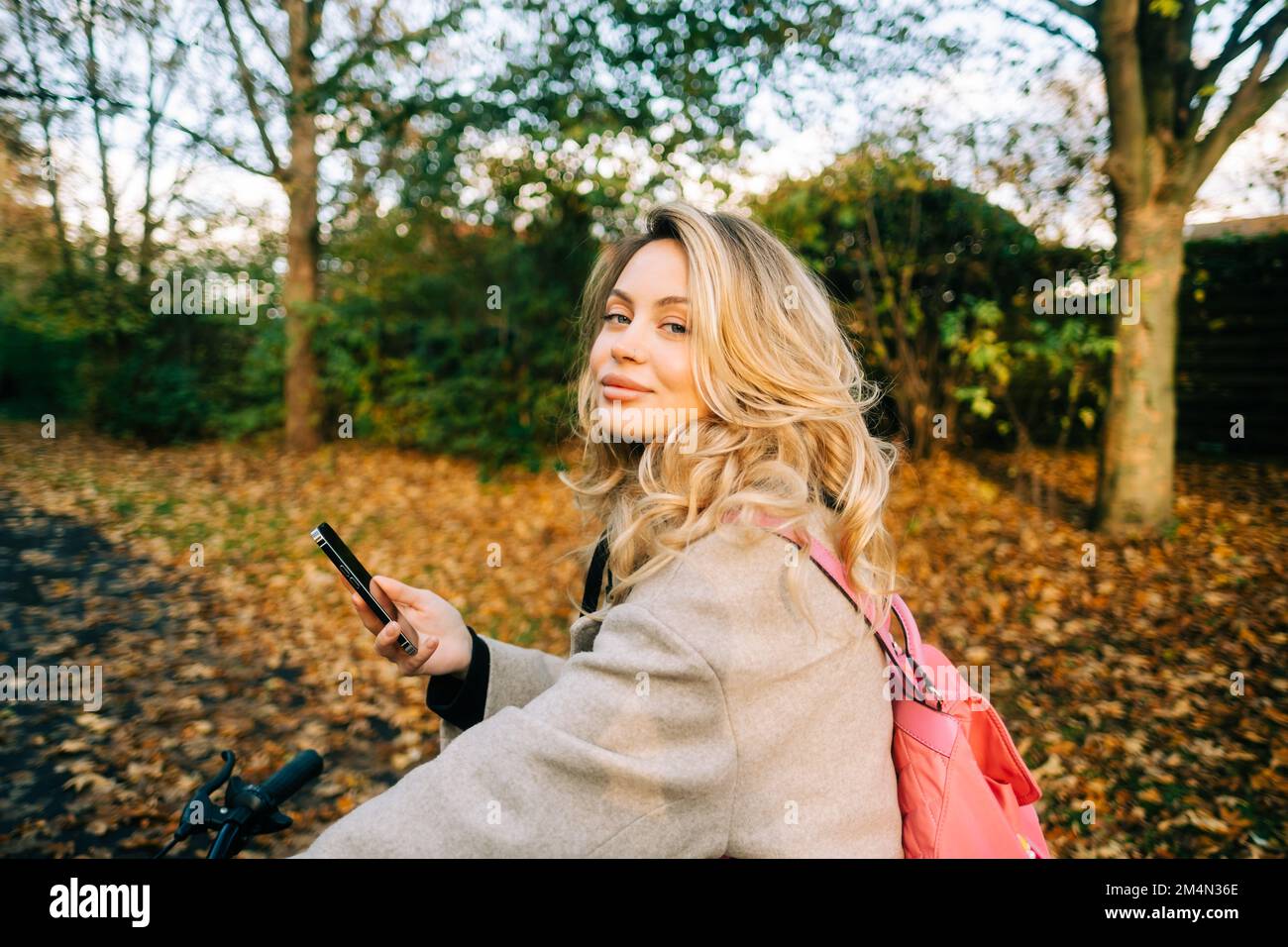 Mujer caucásica atractiva joven que usa el teléfono móvil, paseo en bicicleta en el parque en día cálido en el otoño. Foto de stock