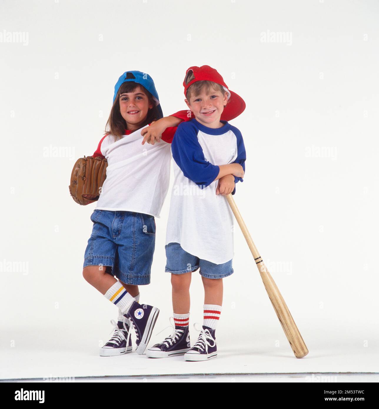 Un niño y una niña vestidos con traje de béisbol con zapatillas de deporte  y sombreros en la muchacha al revés con guante y niño con bate de béisbol  posando para una