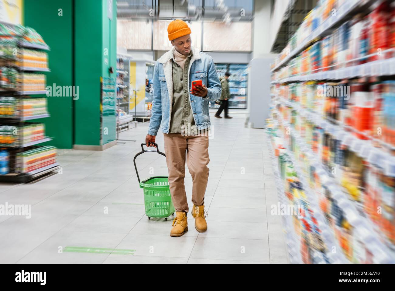 Un tipo negro con estilo con cesta de la compra y smartphone pasa por las estanterías con diferentes productos en el moderno departamento de supermercados Foto de stock