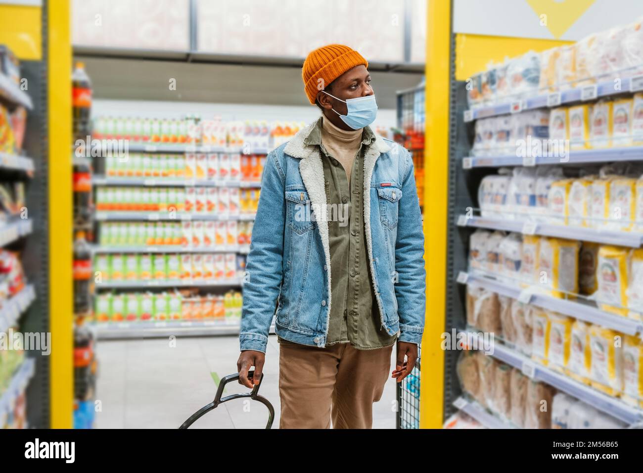 Joven afroamericano con sombrero naranja con máscara protectora camina mirando las estanterías con artículos en el departamento de supermercados Foto de stock