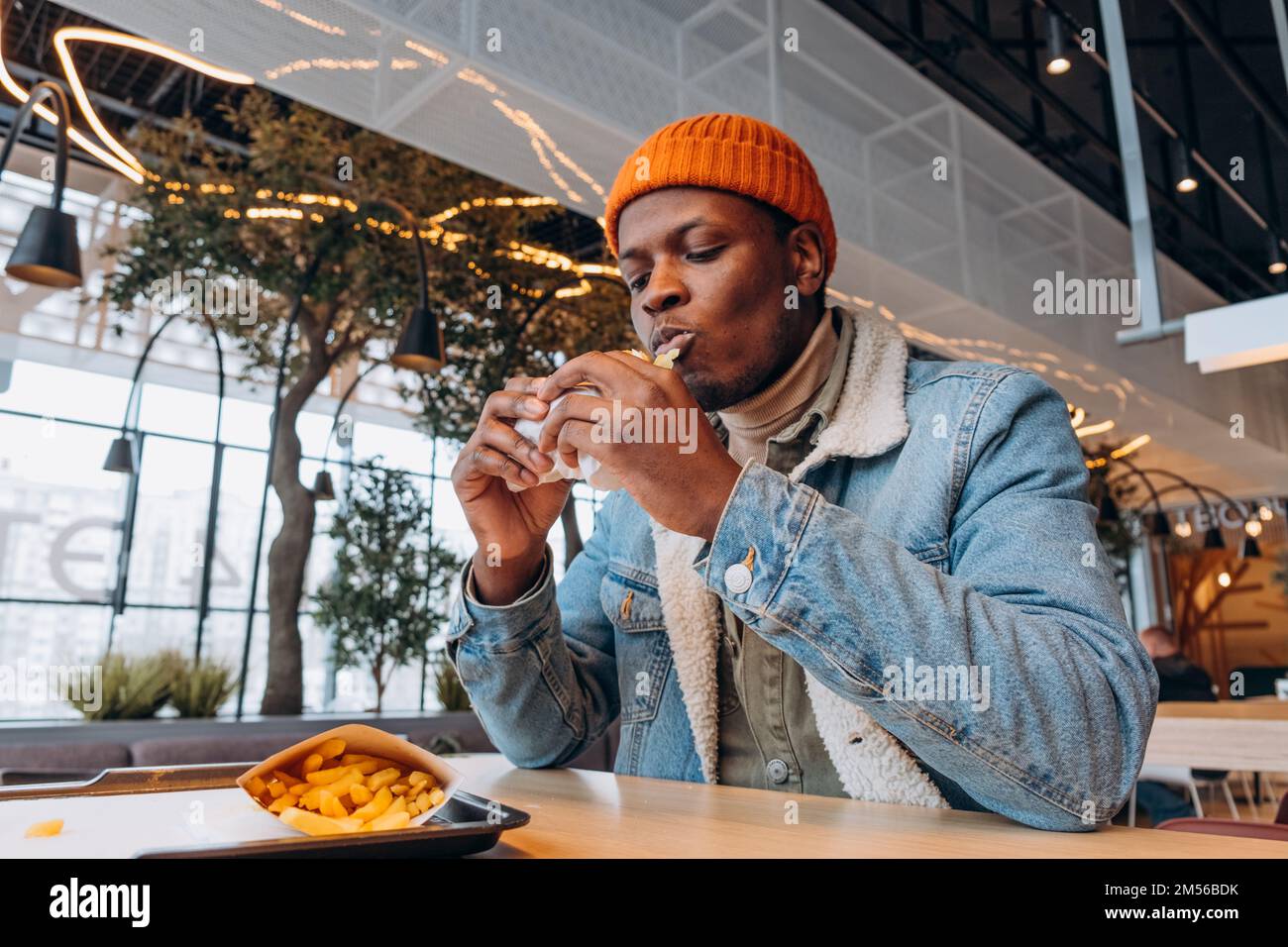 Hombre negro joven en sombrero de punto naranja se come hamburguesa sabrosa en la mesa con papas fritas en la bandeja en el café decorado a la vista Foto de stock