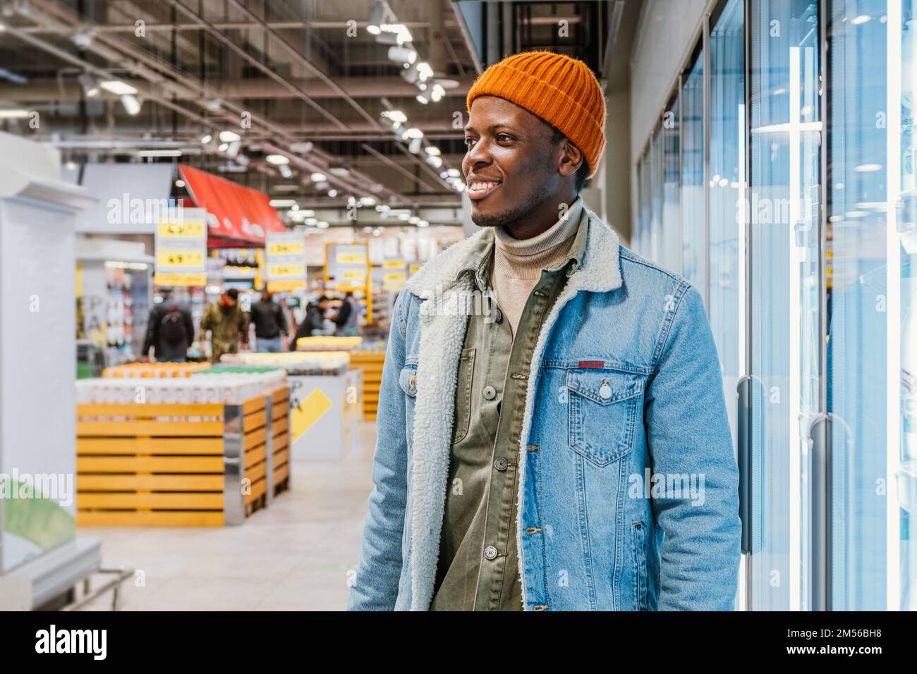 Alegre joven afroamericano con una chaqueta vaquera cálida cerca de los refrigeradores en el moderno departamento de supermercados closeup Foto de stock