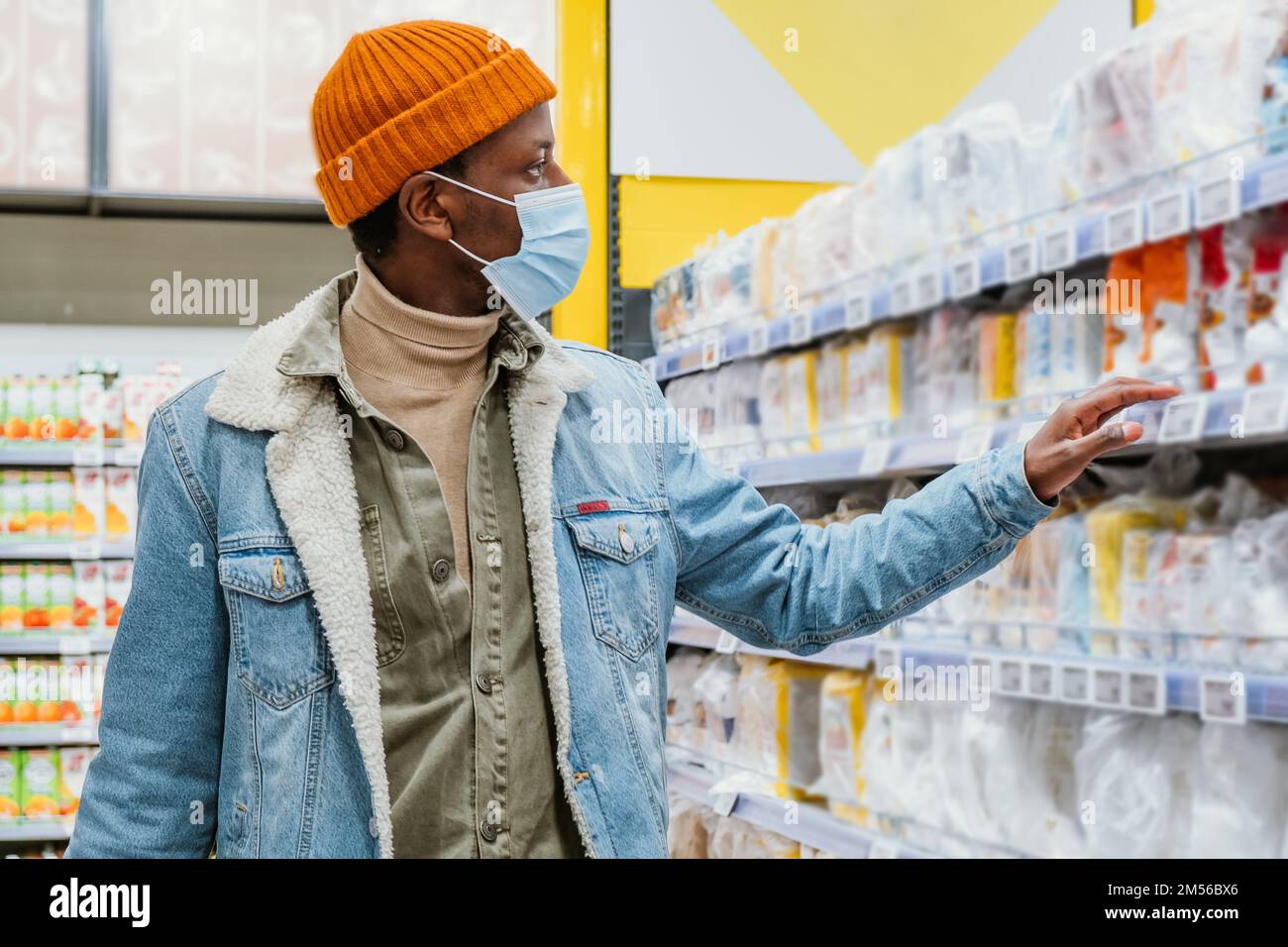 El hombre afroamericano joven en chaqueta de mezclilla con máscara protectora elige mercancías en el estante en el primer plano del departamento moderno del supermercado Foto de stock