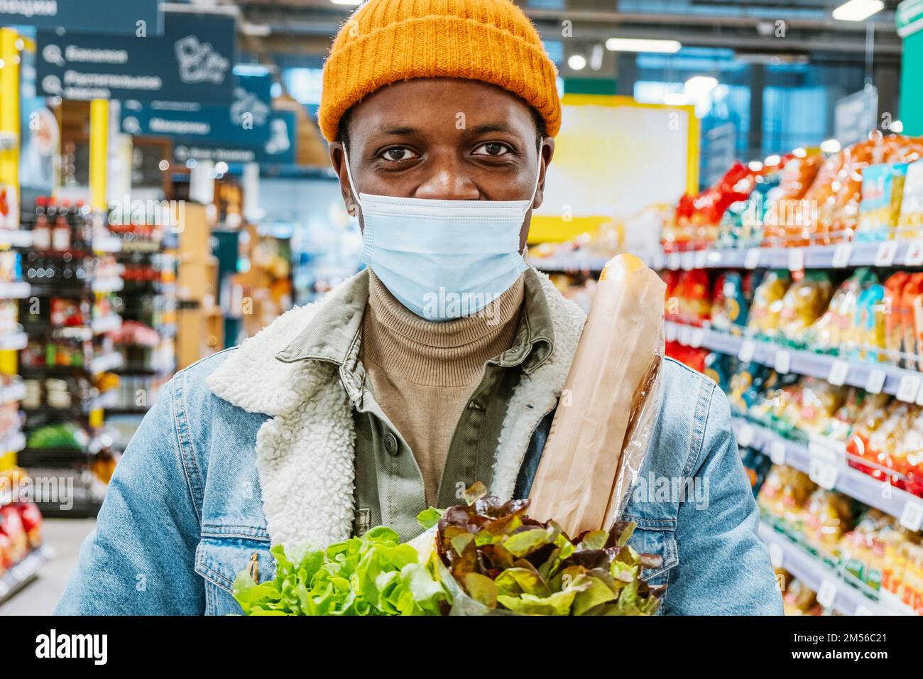 Feliz hombre afroamericano shopper en sombrero naranja de punto con máscara protectora sostiene ensaladas en el departamento de supermercado closeup Foto de stock