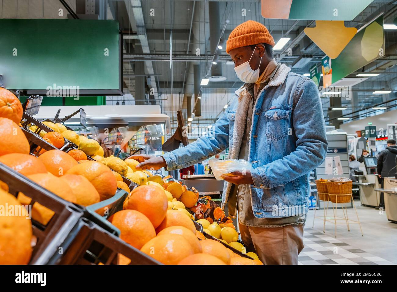 Un hombre afroamericano con chaqueta cálida con máscara desechable saca las frutas frescas de limón de la caja en la vista lateral del supermercado Foto de stock