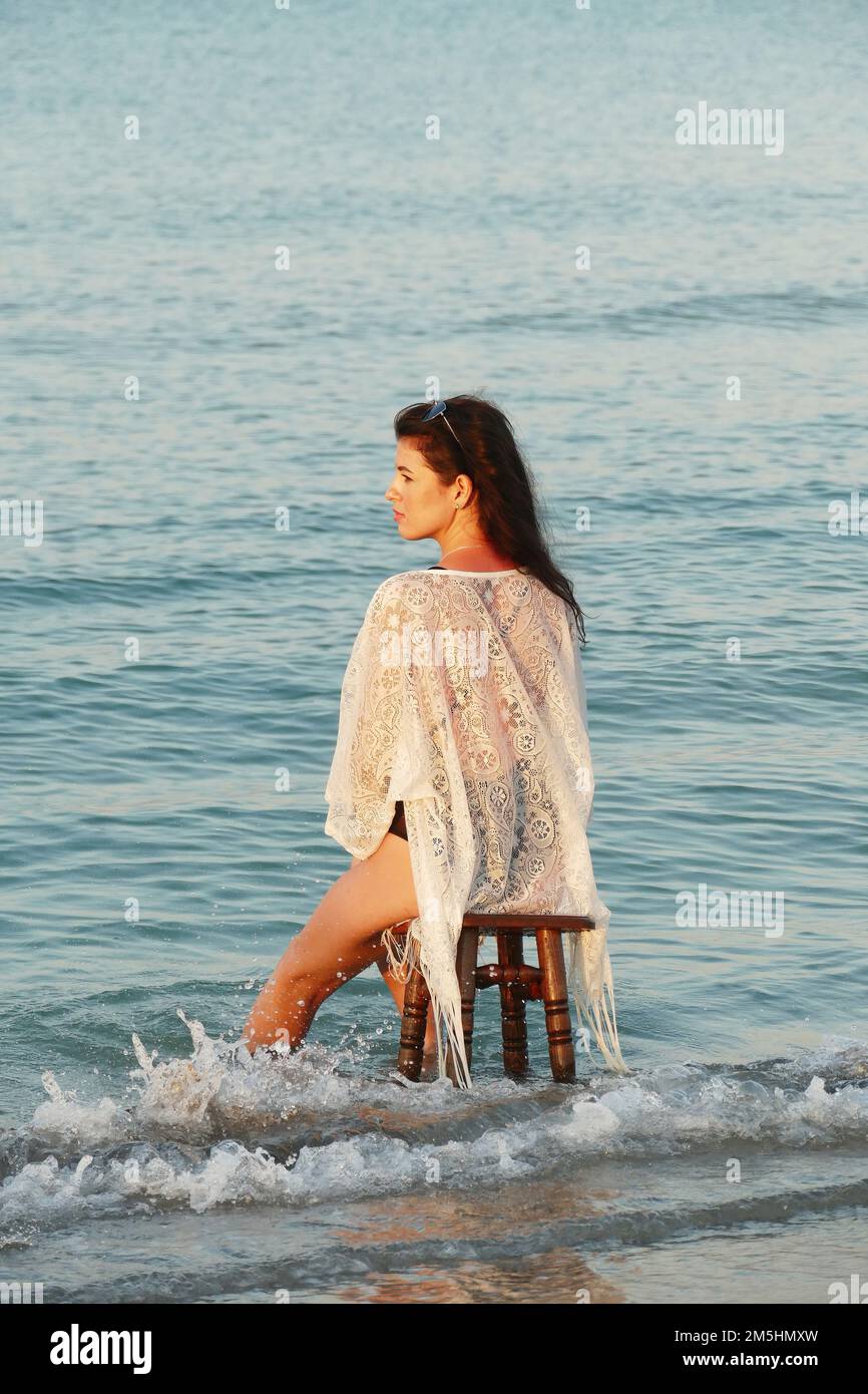 Una mujer hermosa joven posa en la playa del océano. Bastante morena en la playa  blanca túnica y gafas negras sentado en un taburete en el mar Fotografía de  stock - Alamy