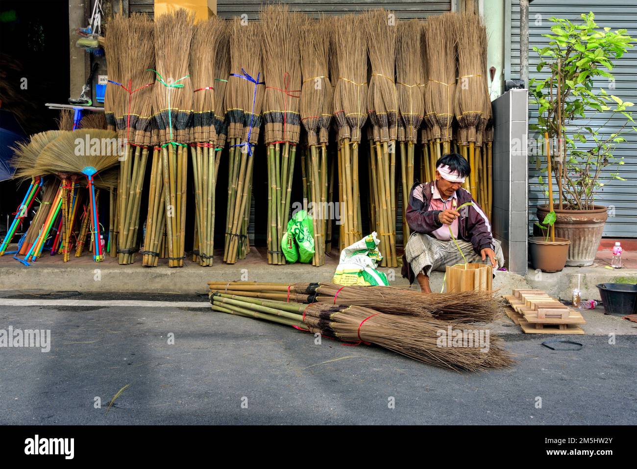 Bangkok, Tailandia, 30 de mayo de 2022: Un trabajador trabaja en un sendero frente a una tienda que vende escobas de tallo de coco y otras herramientas. Foto de stock