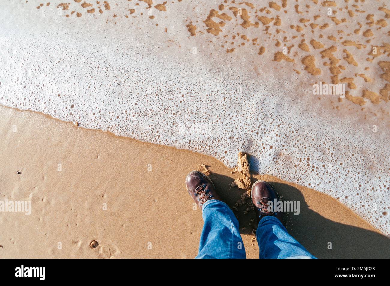 Hombre en jeans y botas de pie en una playa en Francia en diciembre mirando hacia abajo Foto de stock