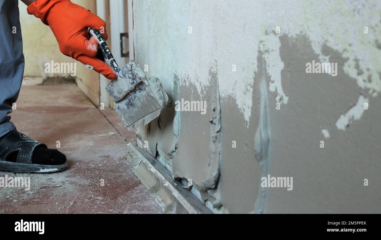 nivelar la pared de una habitación con una gruesa capa de masilla de plástico utilizando una espátula en una mano con guantes de naranja, aplicando mortero de cemento en la superficie de la pared Foto de stock