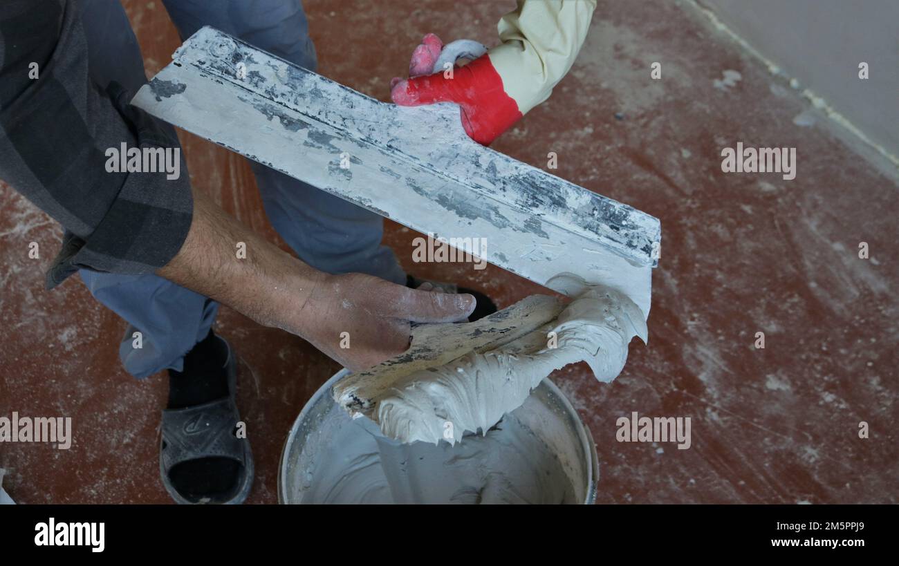 las manos del maestro en un guante sostienen dos paletas con un mortero gris grueso sobre un recipiente, preparando un material de trabajo para el enlucido de las superficies Foto de stock