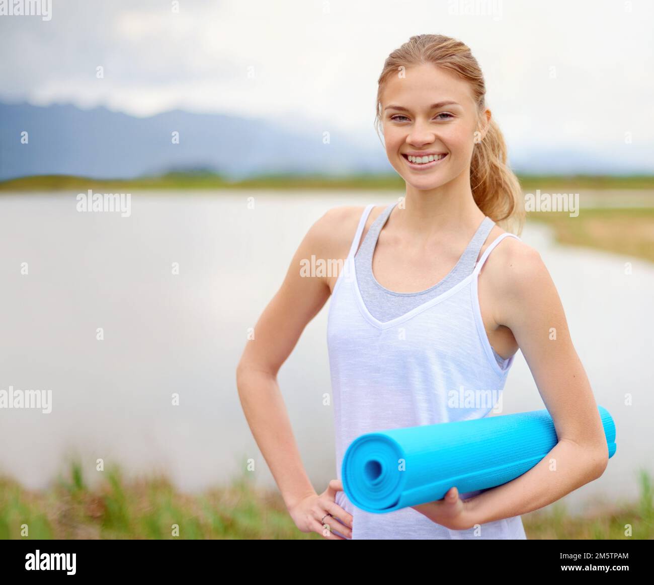 Este parece el lugar perfecto para un poco de yoga. Retrato de una mujer joven feliz haciendo yoga junto a un lago afuera. Foto de stock