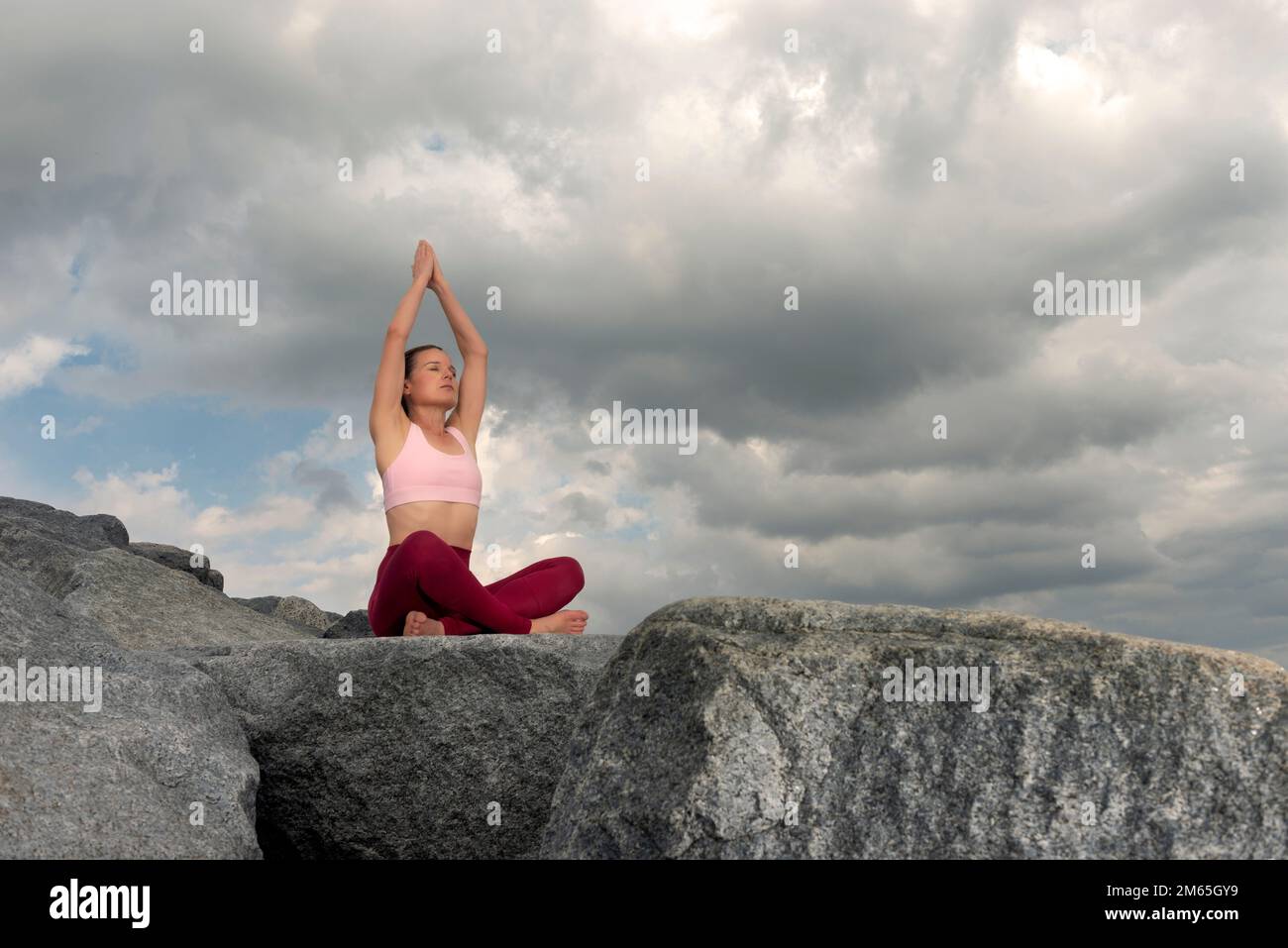 Mujer deportiva sentada con piernas cruzadas en rocas meditando y practicando yoga, cielo dramático. Foto de stock