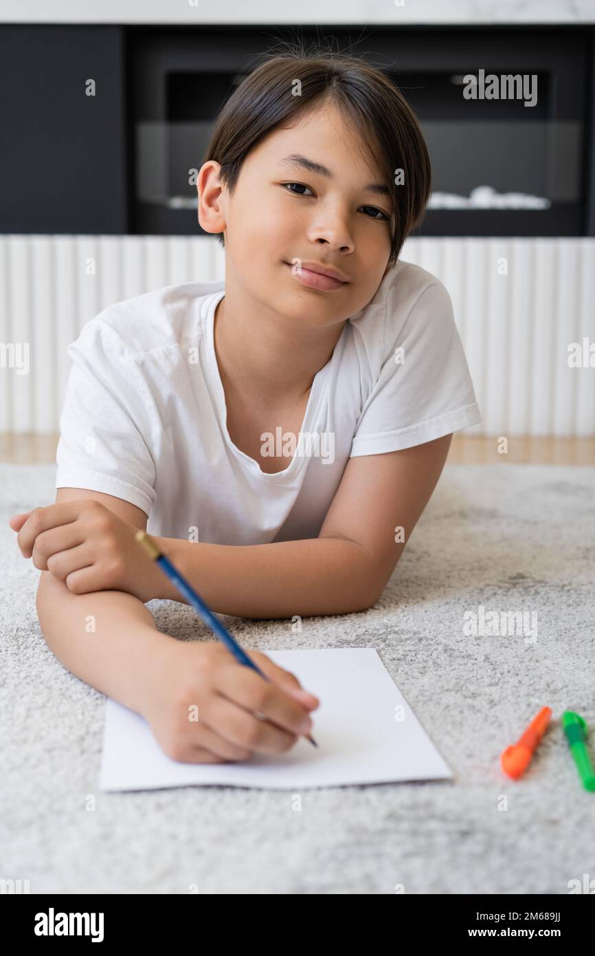 Niño asiático preadolescente mirando a la cámara mientras dibuja en papel en casa, imagen de stock Foto de stock