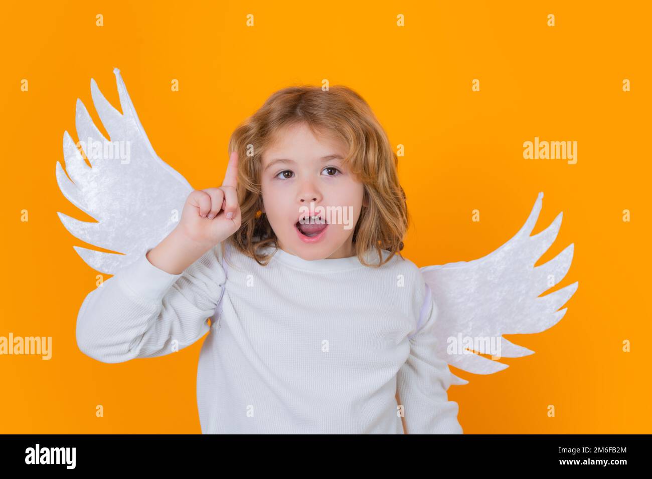 Ángel niño apuntando hacia arriba, gesto de punto, idea. Niños de Navidad. Pequeño cupido ángel niño con alas. Retrato de estudio del niño angelical. Foto de stock