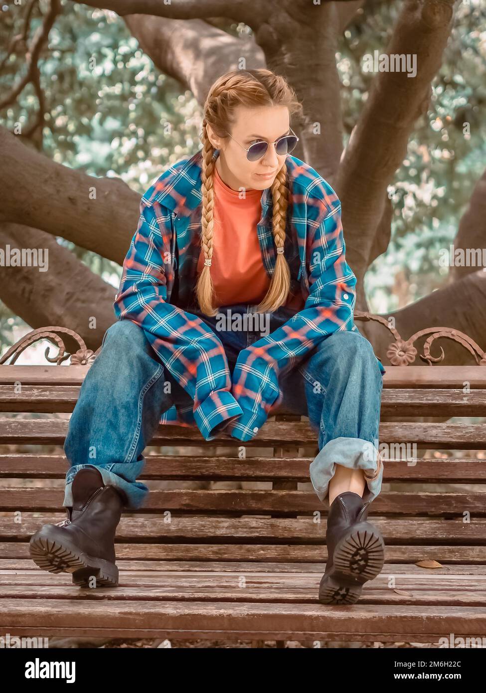 Una chica con coletas en una camisa a cuadros con mangas excesivamente largas, jeans oversize y zapatos ásperos se sienta en un banco del parque. Foto de stock
