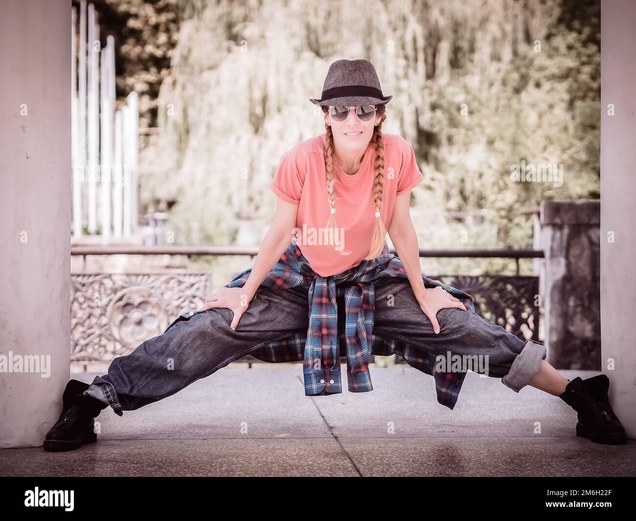 Una chica sonriente con coletas y atuendo casual se coloca con las piernas extendidas entre los pilares del parque. Bailarina callejera. Foto de stock