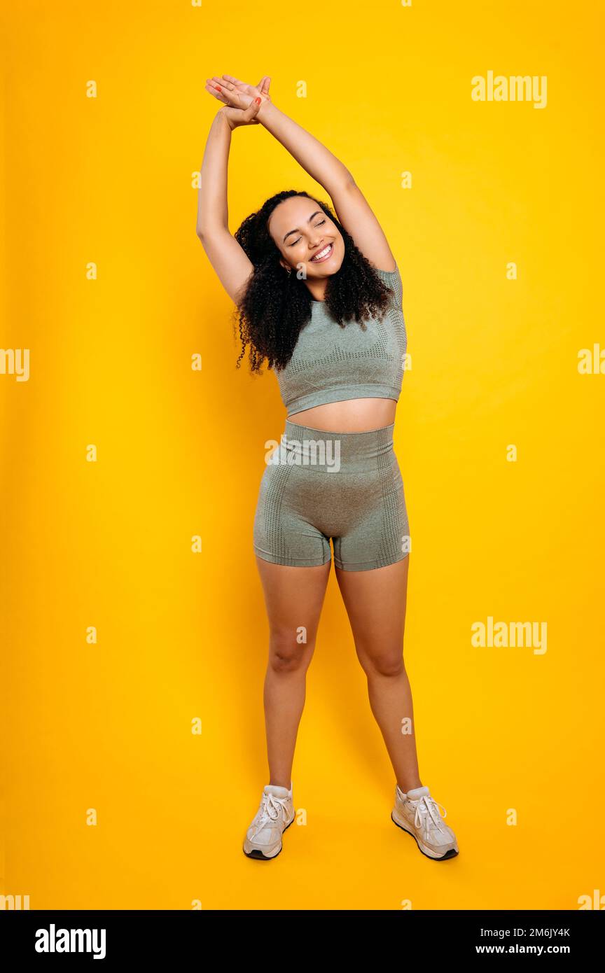Estilo vida deportivo. Foto vertical de longitud completa de mujer brasileña o hispana rizada positiva en ropa deportiva, haciendo estiramiento, estiramiento de los brazos y el cuerpo, para el entrenamiento,