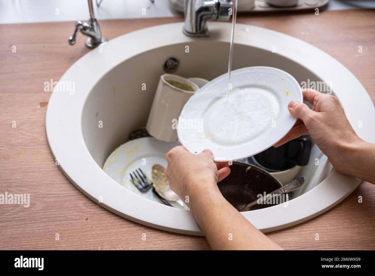 Las manos de las mujeres lavan el plato bajo el agua del grifo. Platos sucios en el fregadero en la cocina, lío después del almuerzo, demasiado perezoso para lavar di Foto de stock