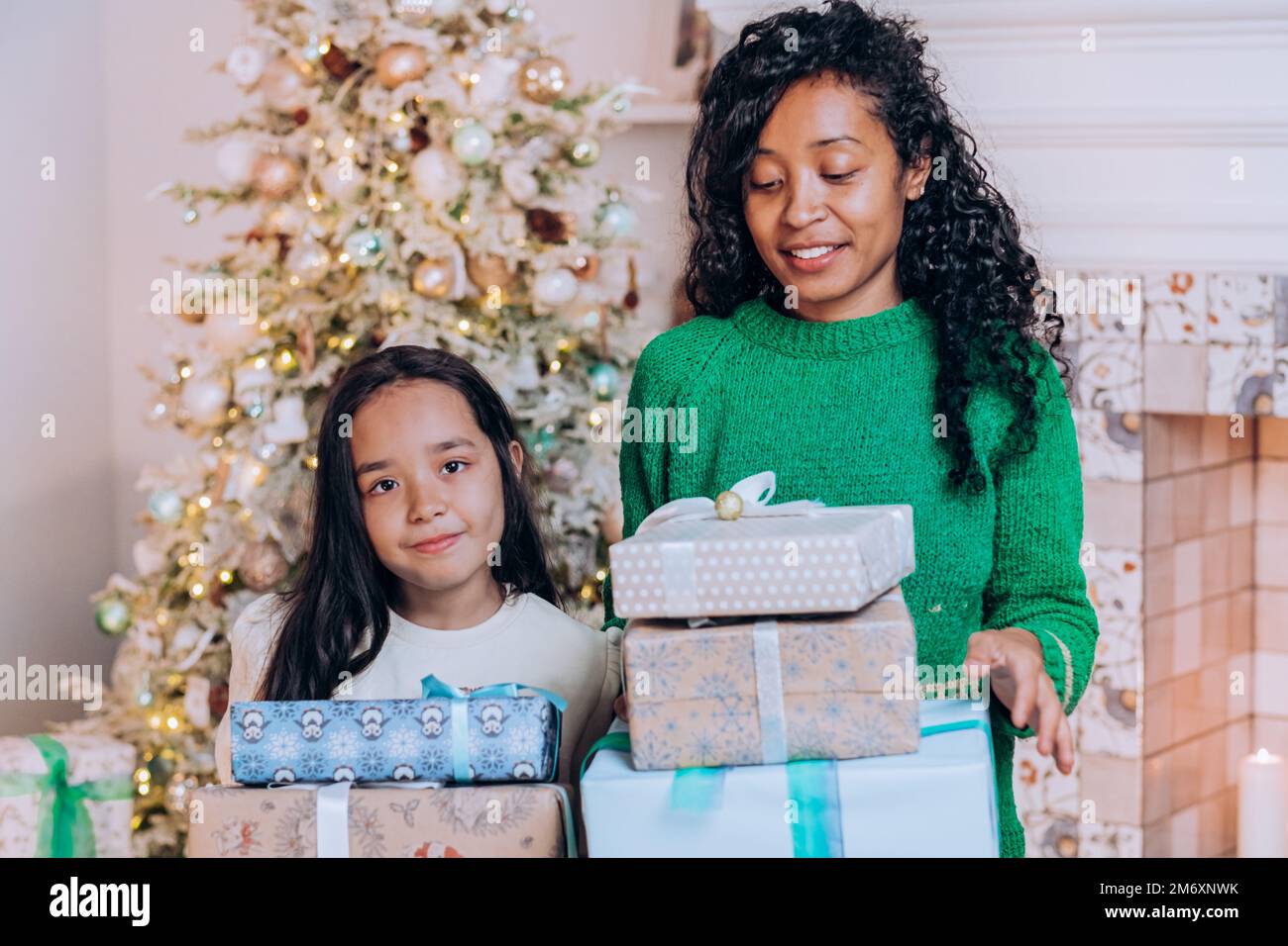 Alegre madre afroamericana e hija morena con pelo largo posan contra decoraciones de árbol de Navidad sosteniendo regalos Foto de stock