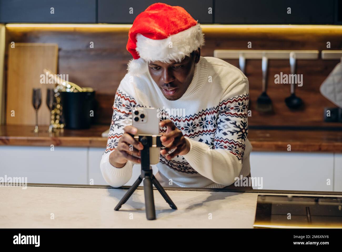 Hombre negro joven en el sombrero de Santa y vídeo impreso caliente de las películas del suéter usando smartphone que se coloca en el trípode en atmósfera de la Navidad en cocina Foto de stock