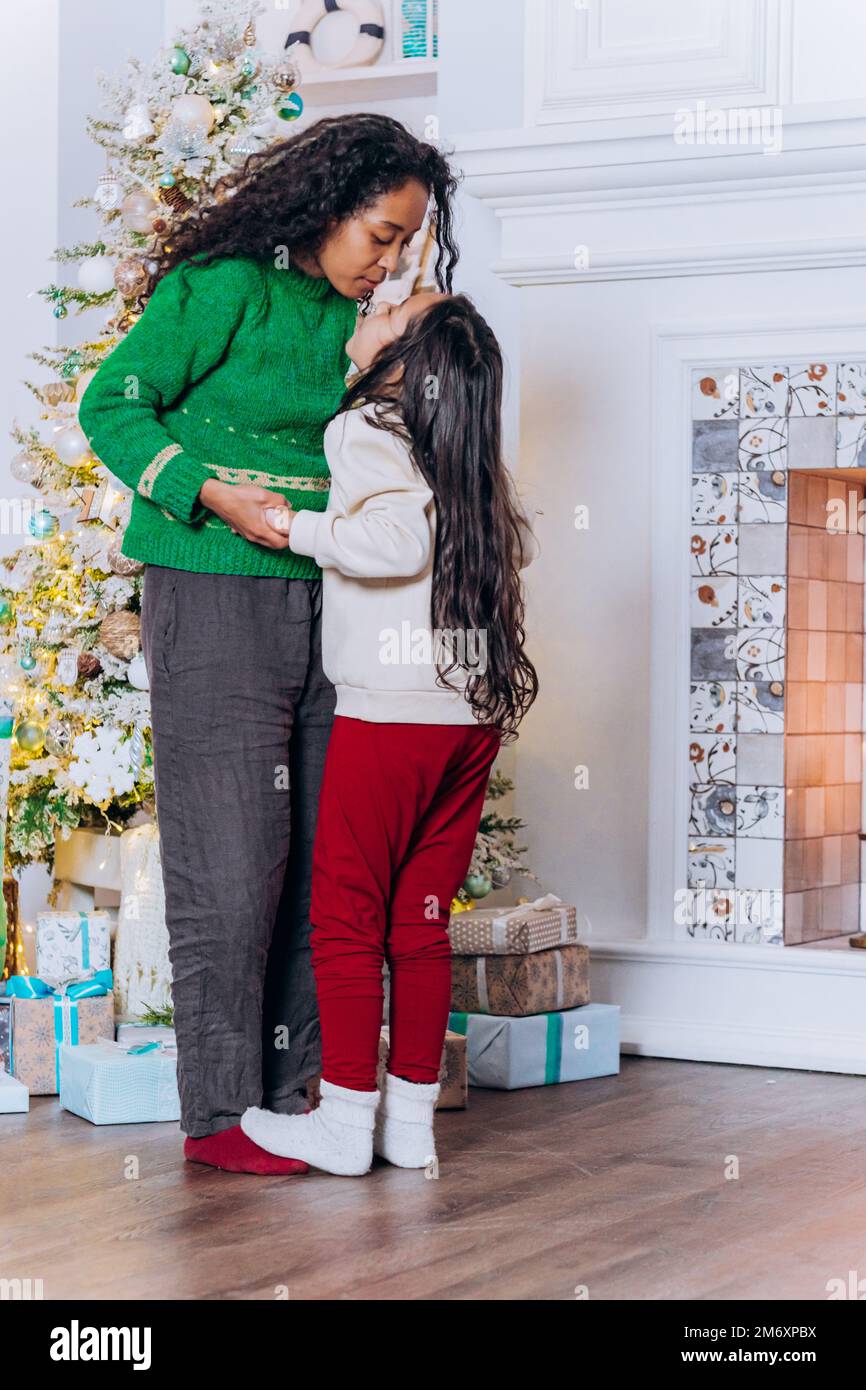 La hija morena de pelo largo juega y baila con la madre afroamericana sonriendo felizmente cerca del árbol de Navidad con cajas de regalo Foto de stock