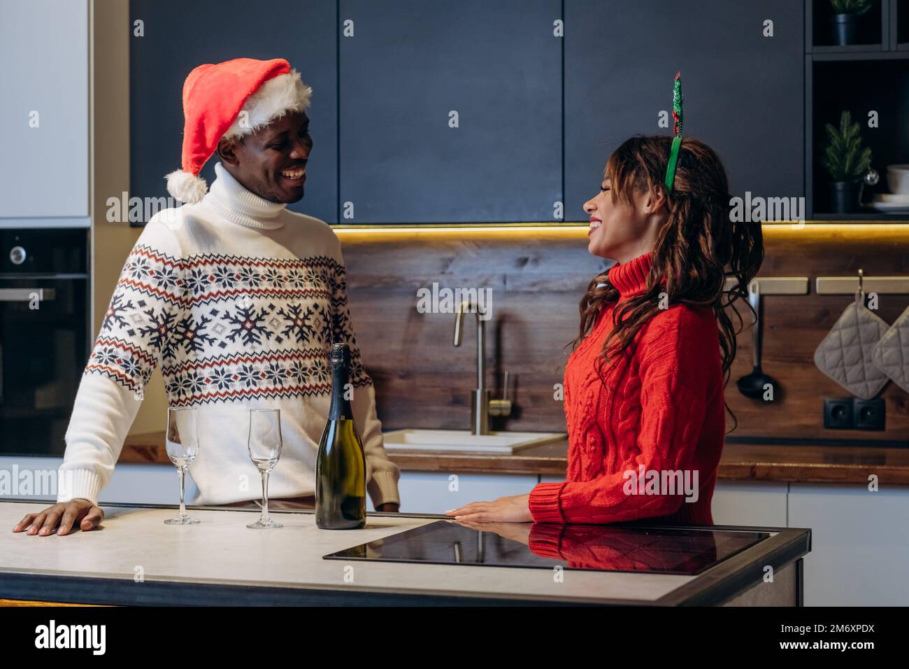 La mujer de pelo rizado y el hombre afroamericano con suéteres impresos festivos beben champán y celebran la Navidad de pie en la cocina Foto de stock