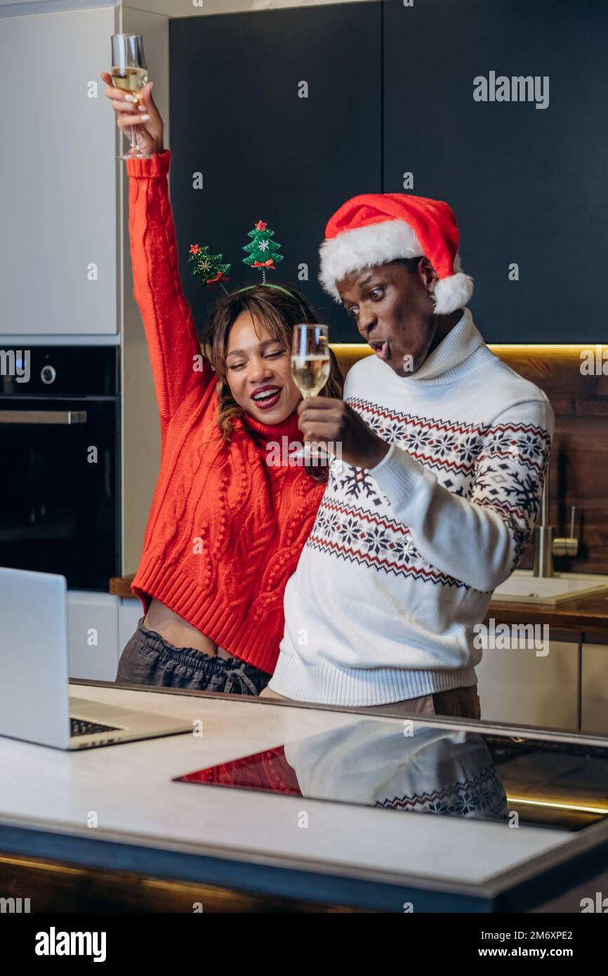 Feliz joven pareja en suéteres festivos y sombreros divertidos habla a través de videollamada con computadora portátil y celebra la Navidad de pie en la cocina Foto de stock