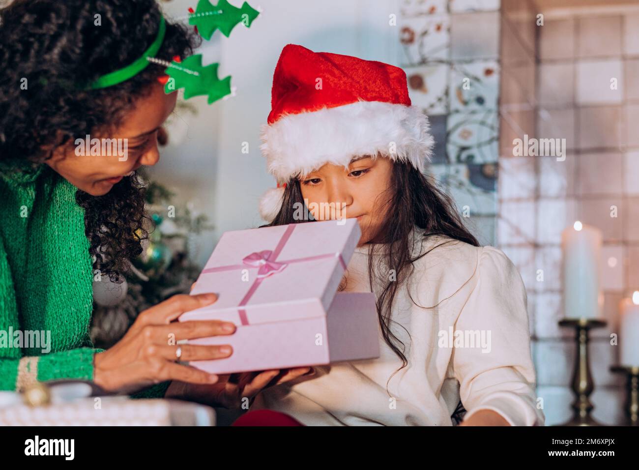 Morena hija de pelo largo en sombrero de Santa abre caja festiva con regalos sentados cerca de la mamá afroamericana contra el árbol de Navidad Foto de stock