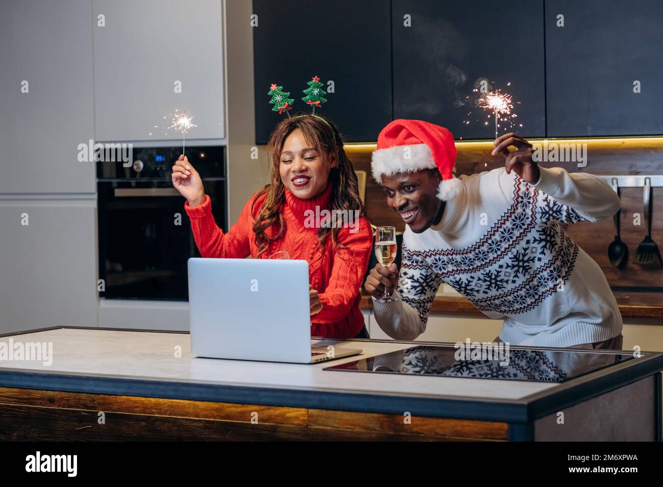 La joven pareja feliz mira los espectáculos festivos usando la computadora portátil y celebra la Navidad sosteniendo chispas y bebiendo champán en la cocina Foto de stock