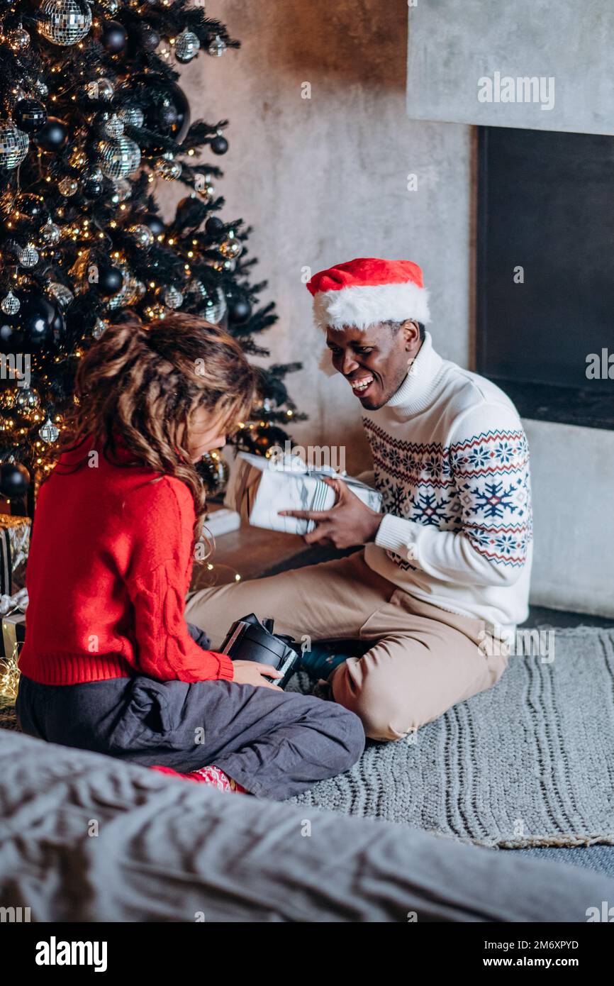 El hombre negro joven en el sombrero de Santa sorprende a la novia morena de pelo largo con el regalo sonriendo brillantemente y sentado contra el árbol de Navidad Foto de stock
