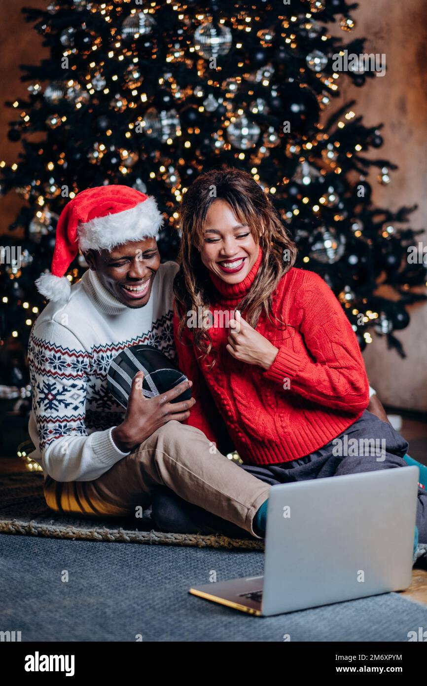 Pareja en suéteres festivos habla a través de videollamada con computadora portátil y sostiene presente sentado contra el árbol de Navidad con adornos y guirnalda Foto de stock