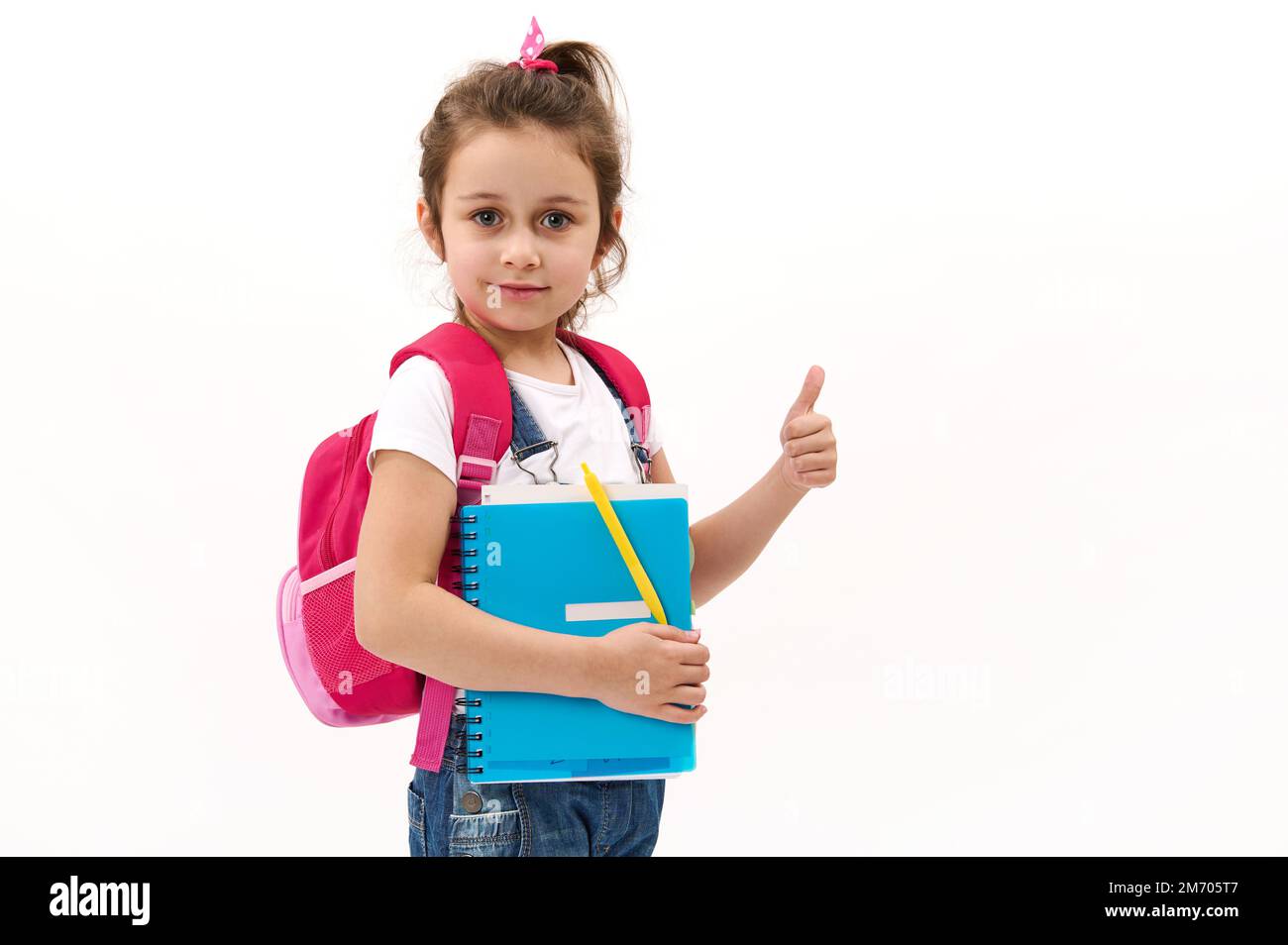 Adorable niña de 5-6 años con mochila rosa, sosteniendo libro de texto y  mostrando el pulgar hacia arriba mirando a la cámara. Maqueta Fotografía de  stock - Alamy