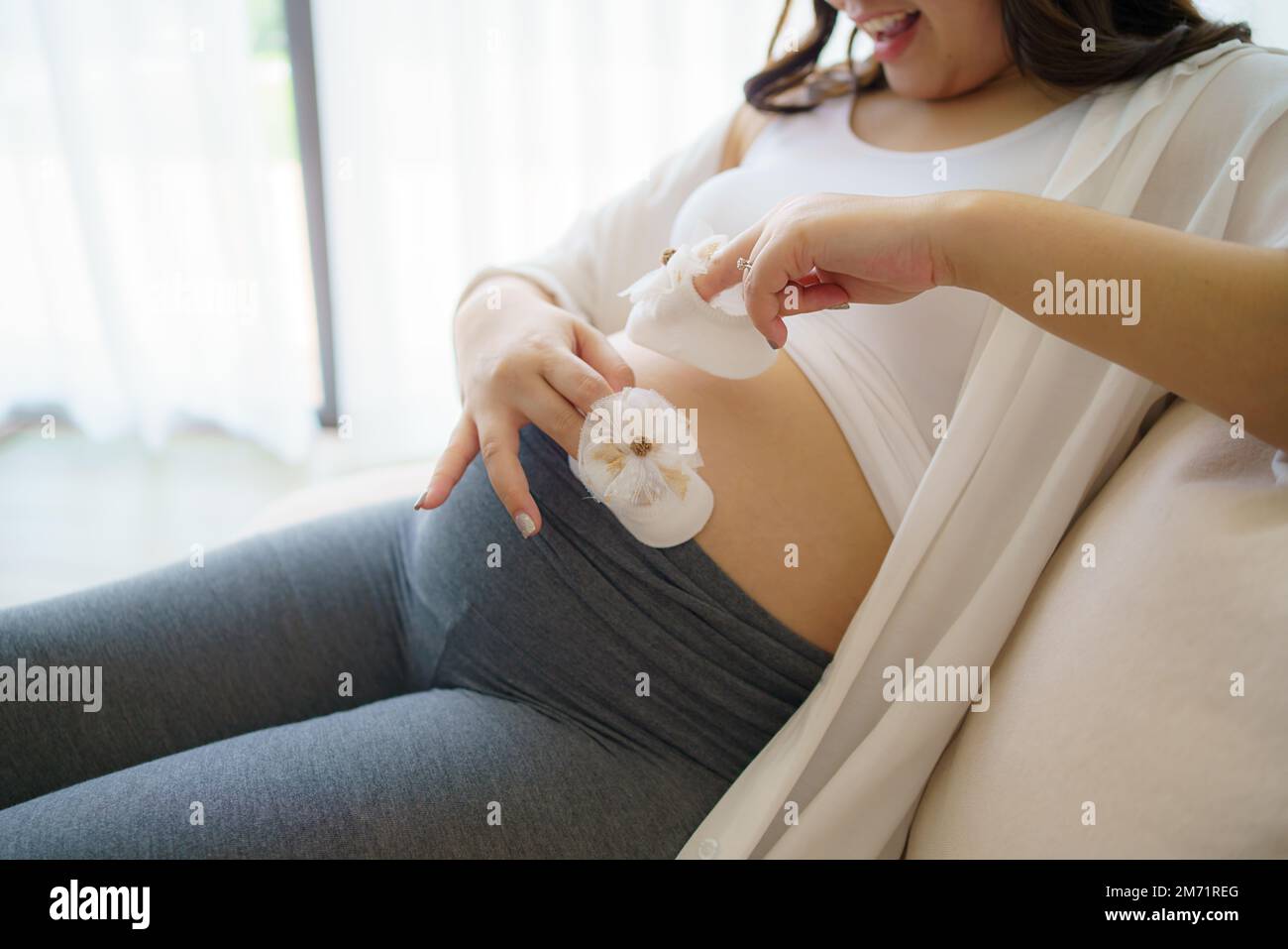 Mujer asiática embarazada que se siente feliz en casa mientras que cuida de su hijo. La joven madre que espera que sostiene al bebé en el vientre embarazada. Maternidad prenat Foto de stock