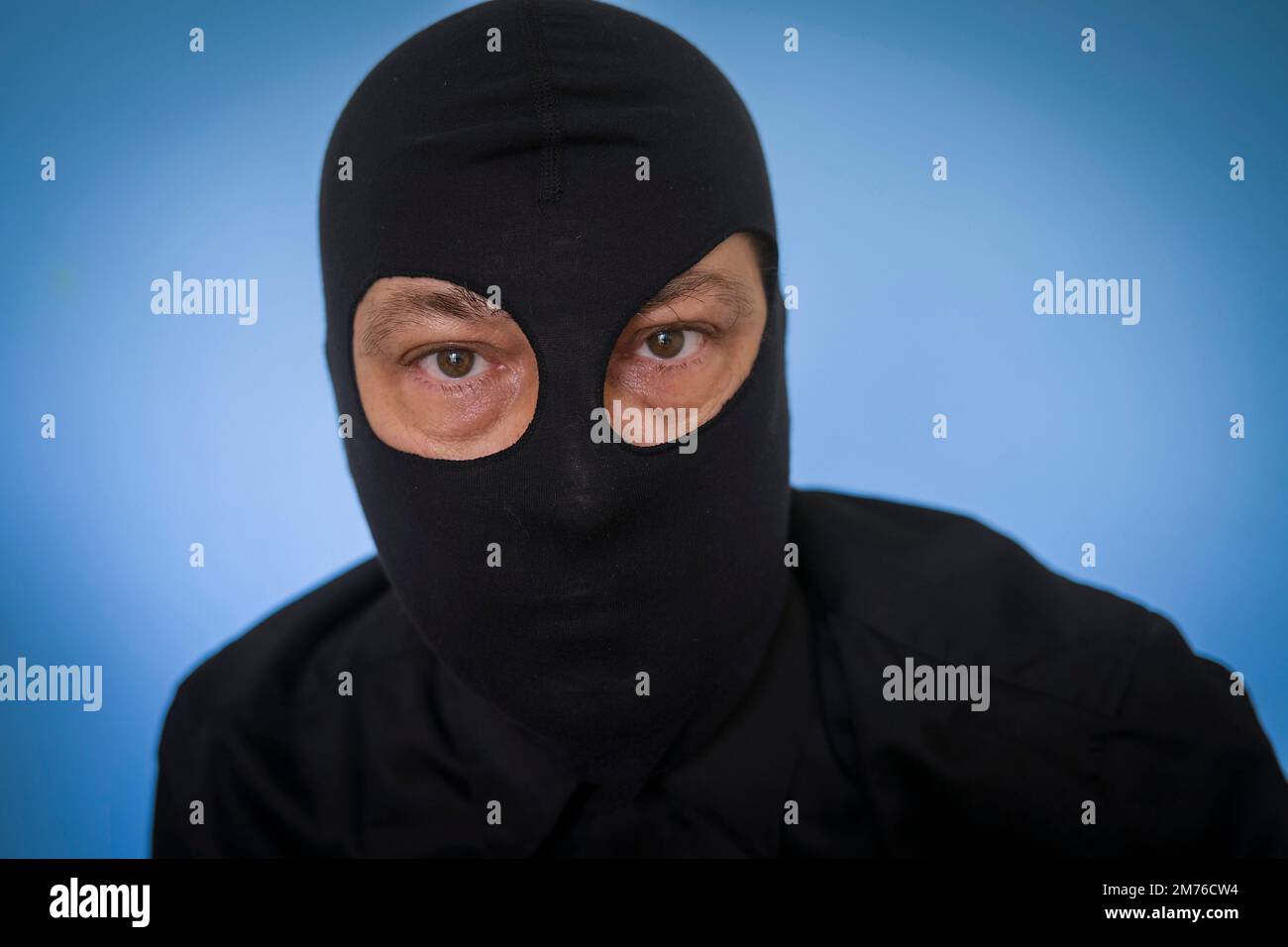 hombre con camisa negra y balaclava negro contra fondo azul, highjacker, terrorista, ladrón, ladrón, concepto de miedos Foto de stock