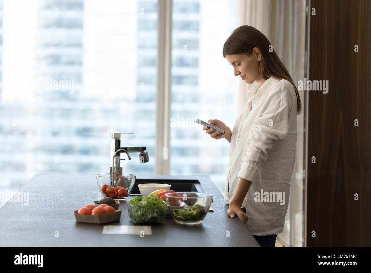Mujer joven que usa smartphone en la cocina mientras cocina ensalada vegetariana Foto de stock