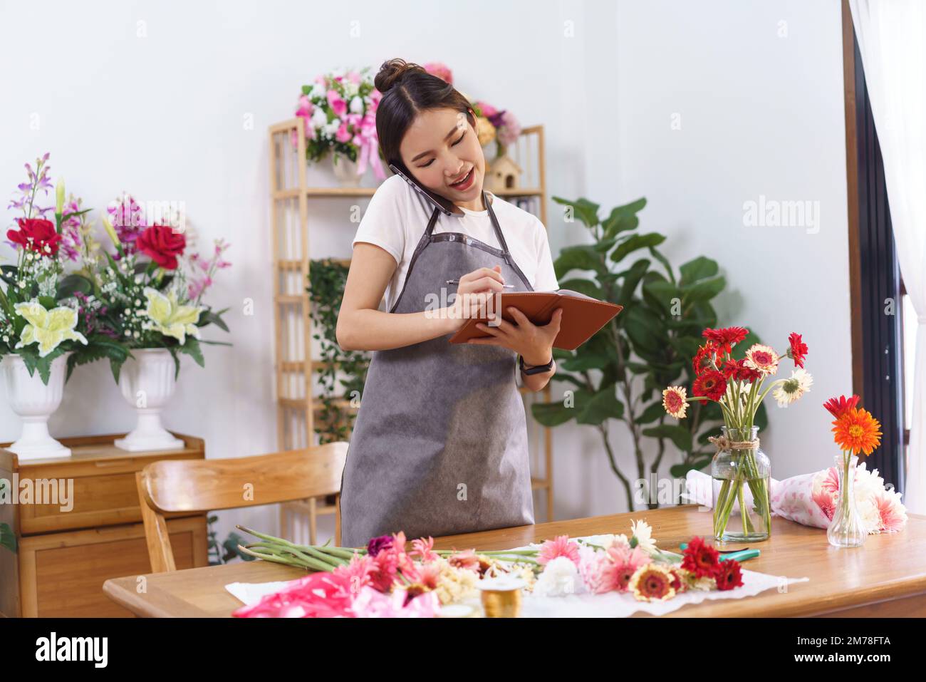 Concepto de tienda de flores, florista femenino sostiene el teléfono entre la cara y el hombro para hablar con el cliente. Foto de stock