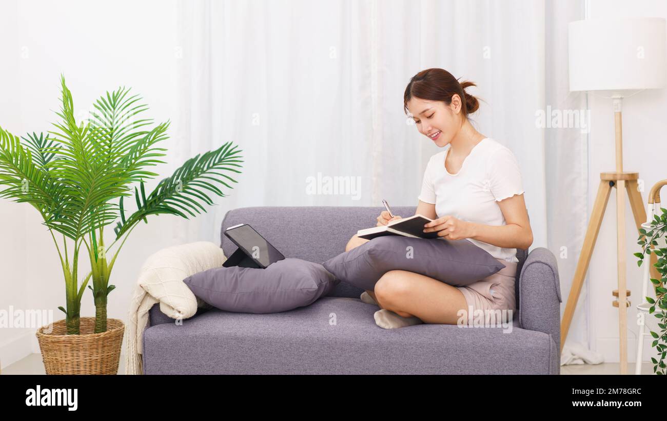 Estilo de vida en el concepto de sala de estar, mujer asiática joven escribiendo datos en el cuaderno mientras se sienta en el sofá. Foto de stock