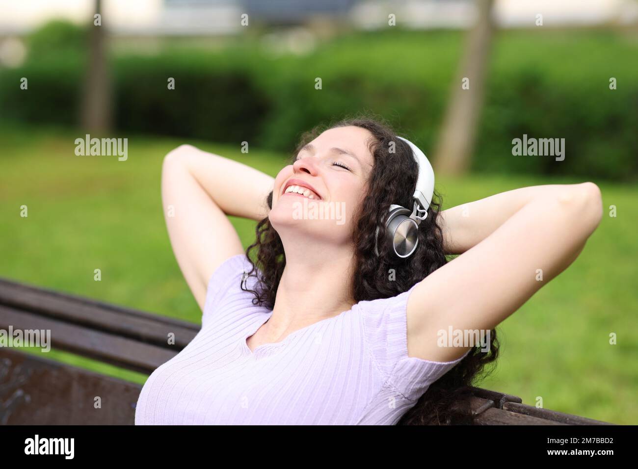 Mujer feliz que descansa sentada en un banco que escucha la música en un parque Foto de stock