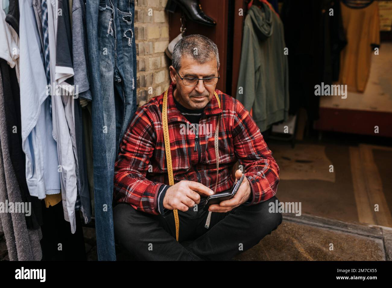 Dueño masculino maduro usando anteojos usando teléfono inteligente mientras se sienta fuera de la tienda de ropa Foto de stock
