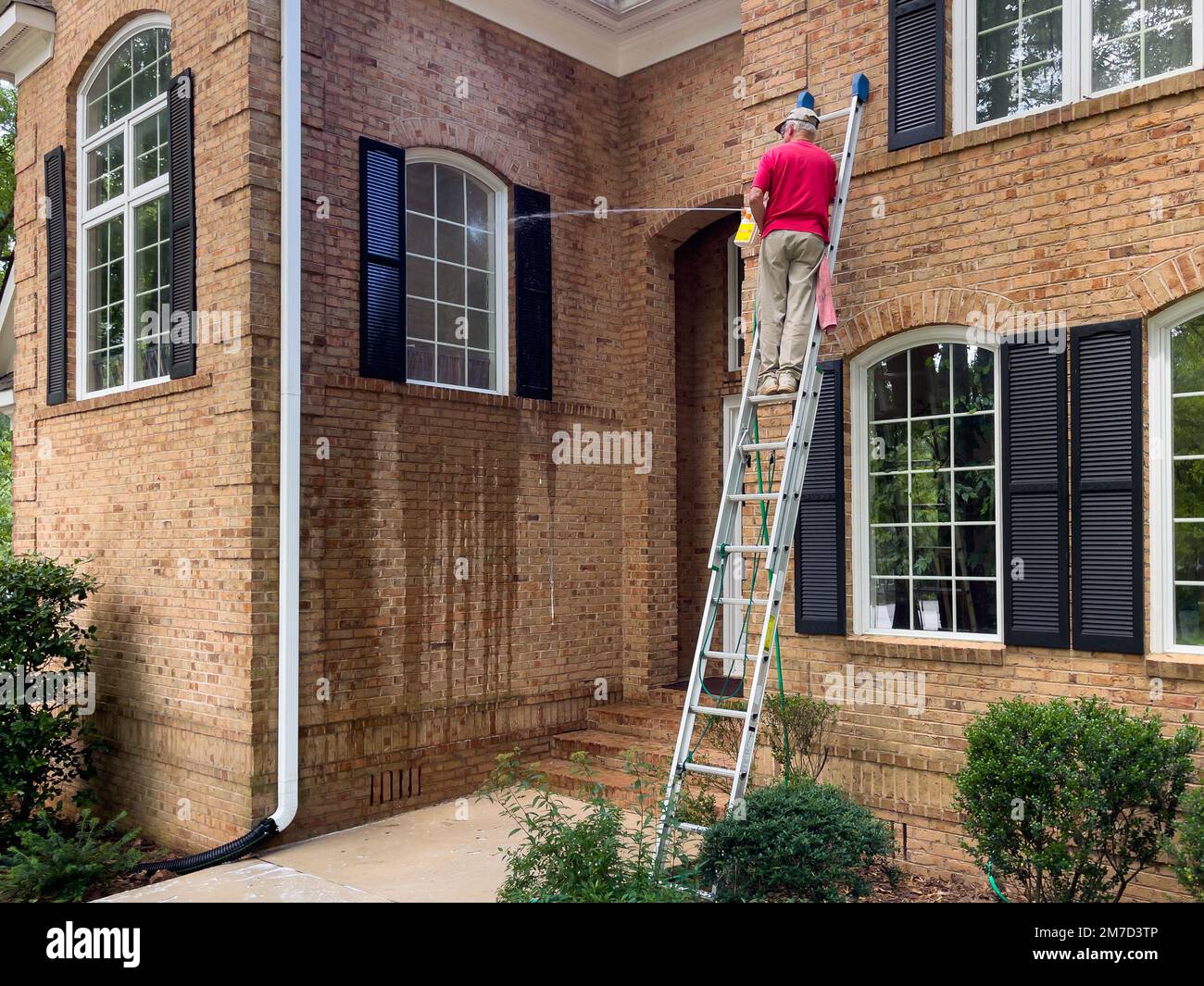 Hombre en escalera lavando ventanas de una casa con un rociador de jabón de lavado a presión. Limpieza del muelle con manguera y arandela a presión. Servicio de limpieza. Foto de stock