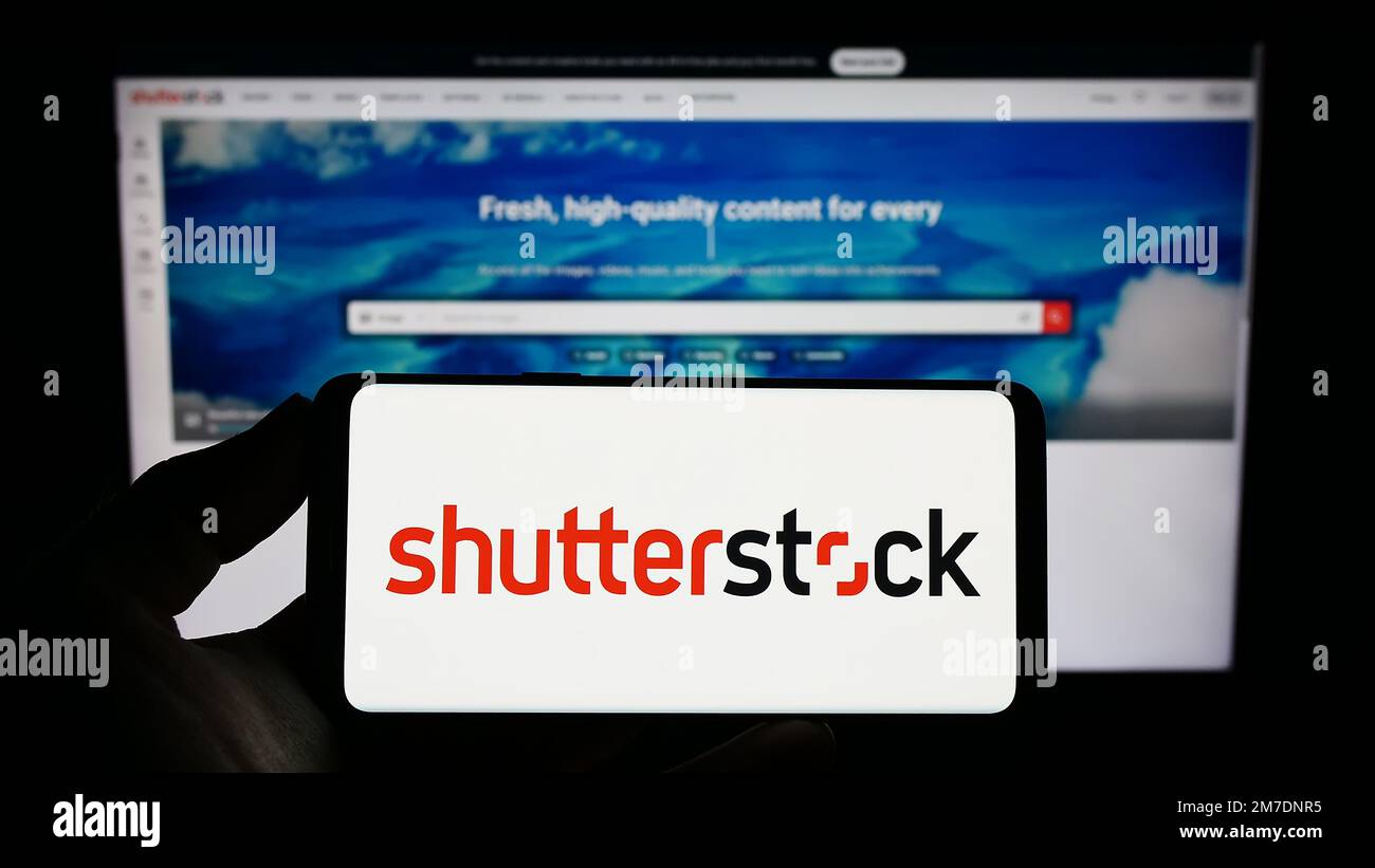 Persona que sostiene el teléfono inteligente con el logotipo de la compañía estadounidense de fotografía de stock Shutterstock Inc. En la pantalla frente al sitio web. Enfoque en la pantalla del teléfono. Foto de stock